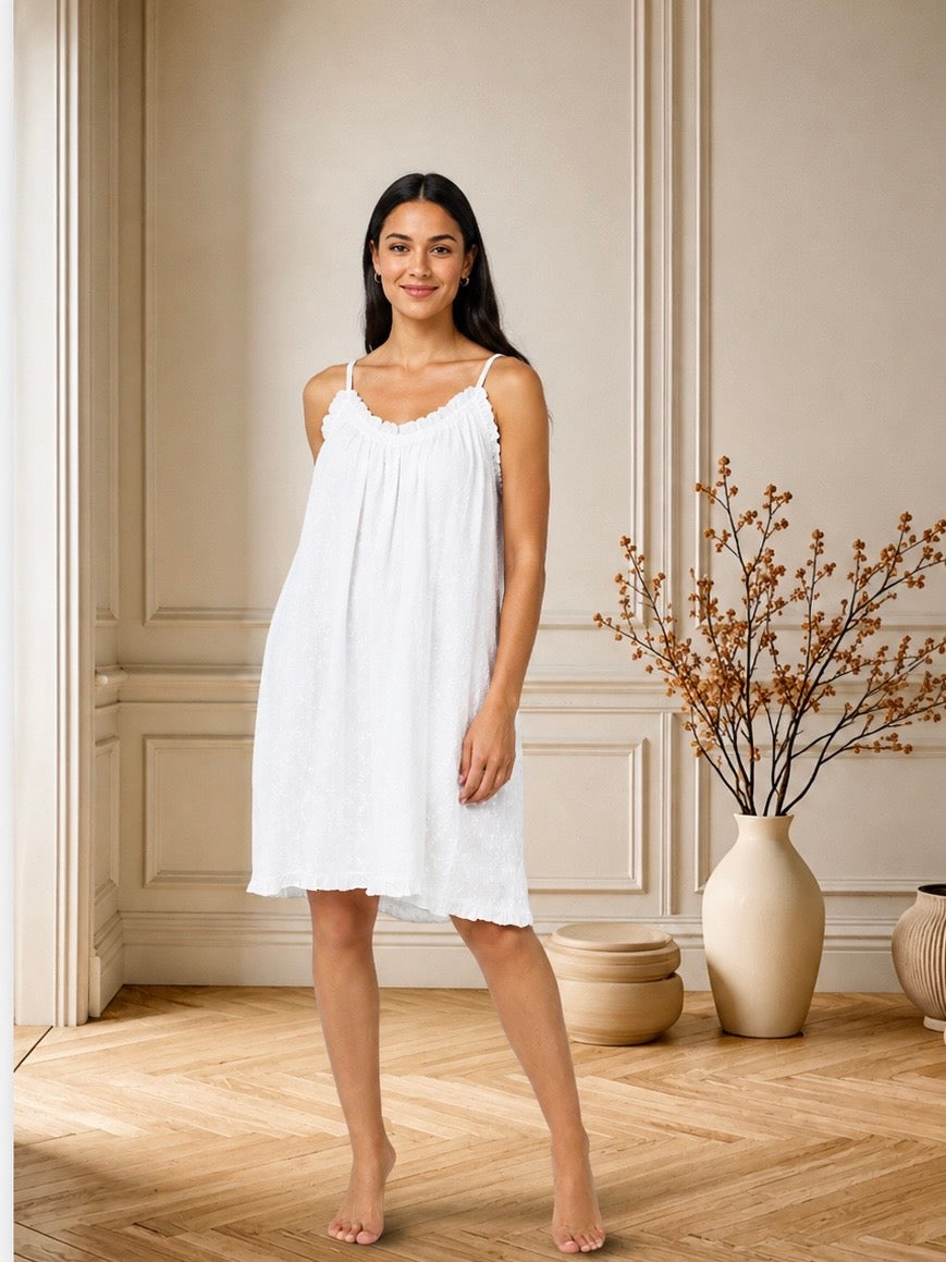 A woman modeling a white sleeveless nightgown with thin straps and a scalloped neckline, standing barefoot in a warm-toned room with paneled walls, a wood floor, and a vase of dried branches.