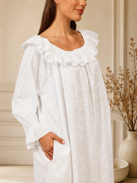 A woman wearing a white long-sleeve nightgown with a ruffled neckline and a subtle textured pattern, standing in a softly lit indoor setting.