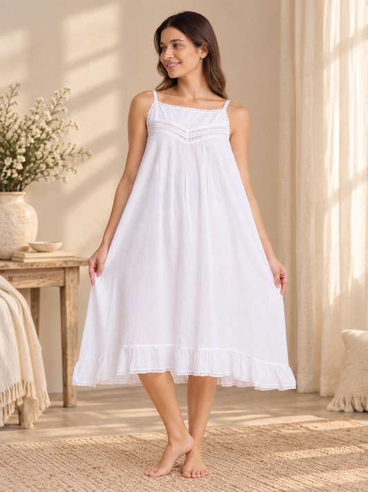 A woman wearing a white sleeveless nightgown with thin straps and a ruffled hem, standing barefoot in a softly lit bedroom setting with curtains, a side table, and a vase of flowers.