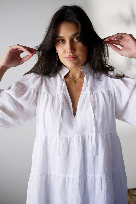 Woman wearing crisp white linen layered dress with tiered panels, voluminous sleeve and neck tie
