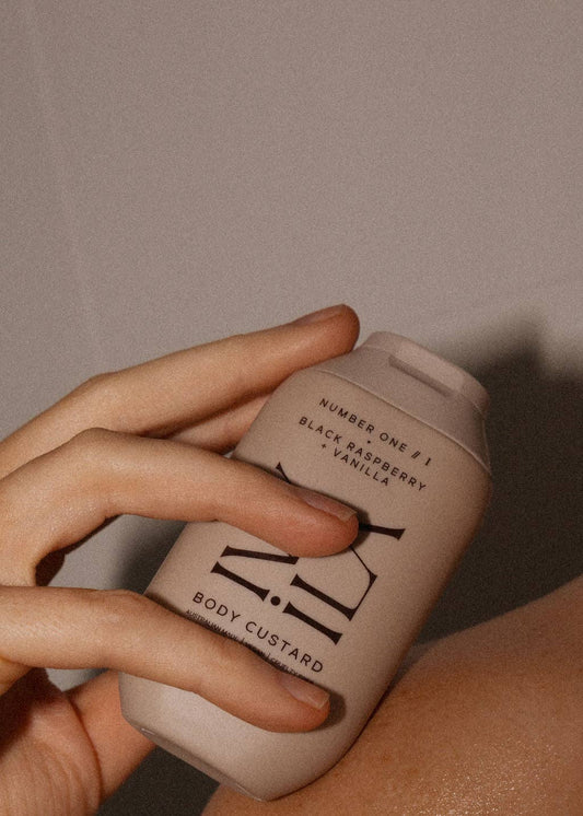 Hand holding a beige bottle labeled 'Body Custard' against a neutral background