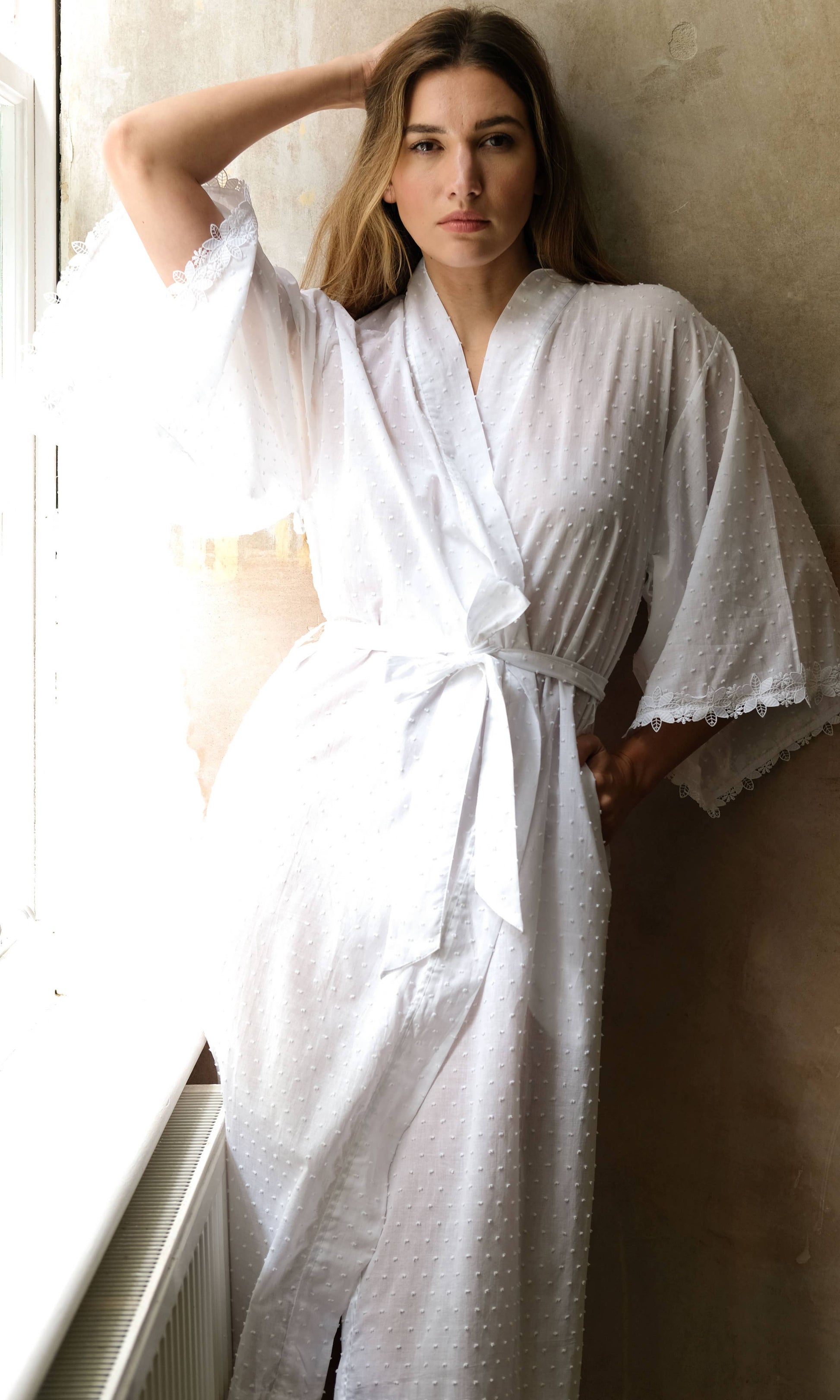 Woman wearing a white robe standing by a window.