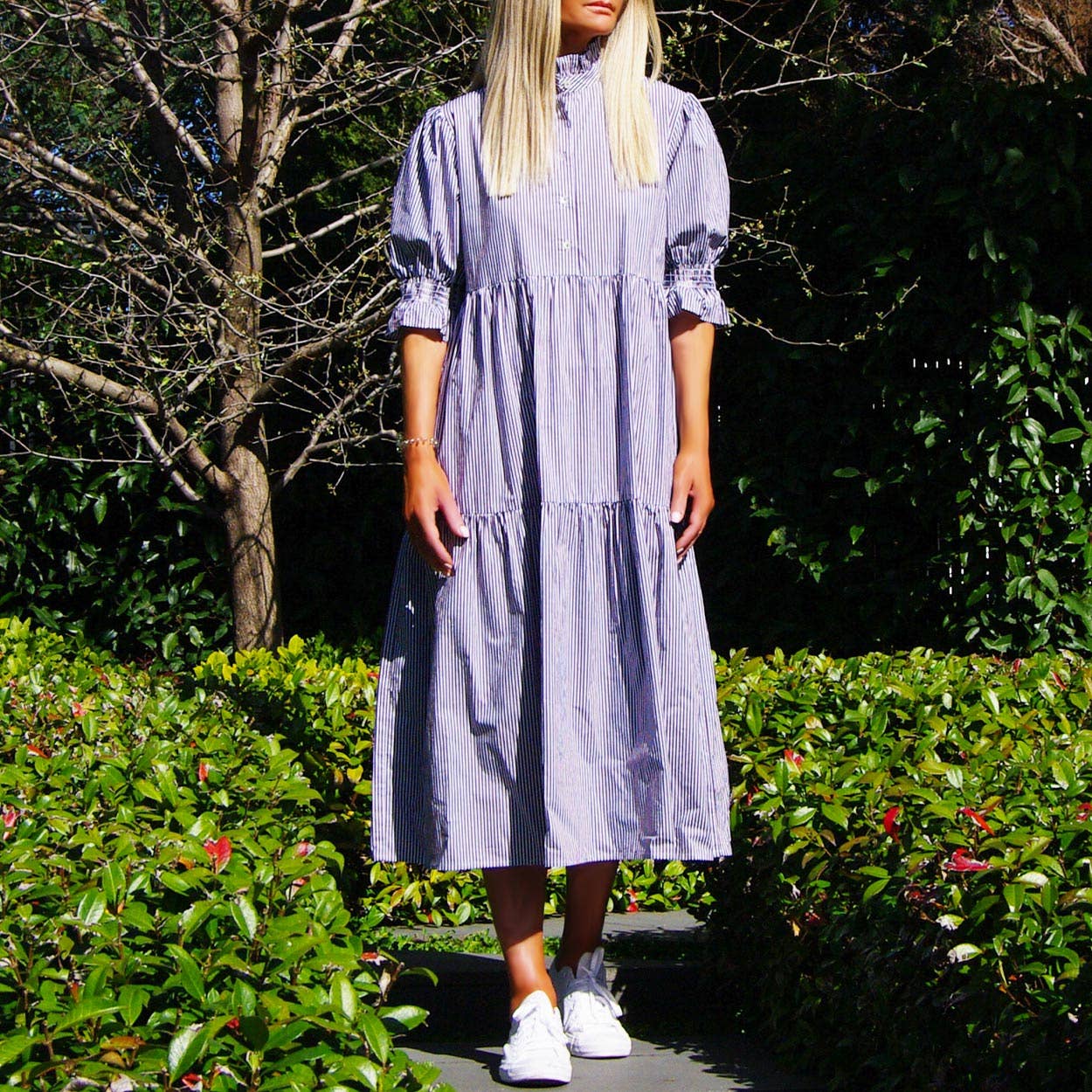 Woman wearing a blue dress standing outdoors with greenery in the background
