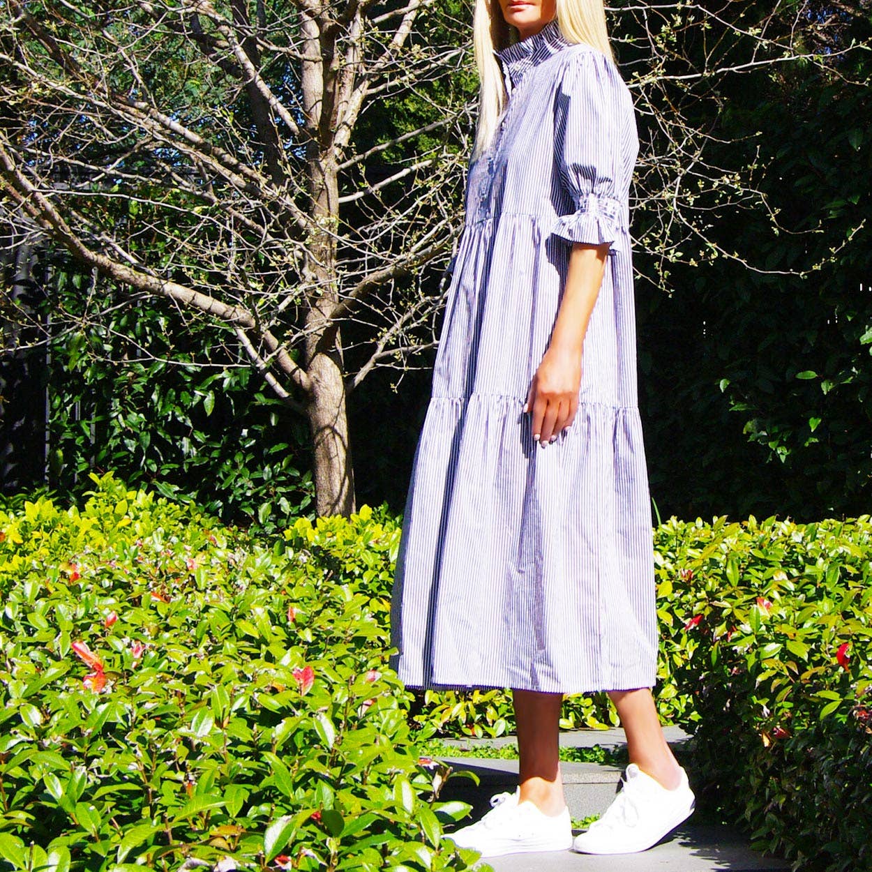 Woman wearing a blue pinstripe dress standing in a garden with greenery and trees in the background.
