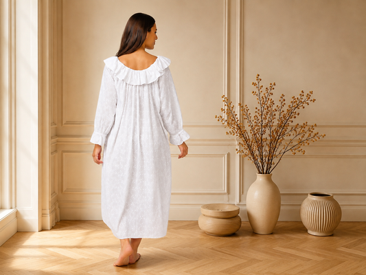 A woman wearing a white long-sleeve nightgown with a ruffled collar, standing in a warm beige room with wood floors and decorative vases.