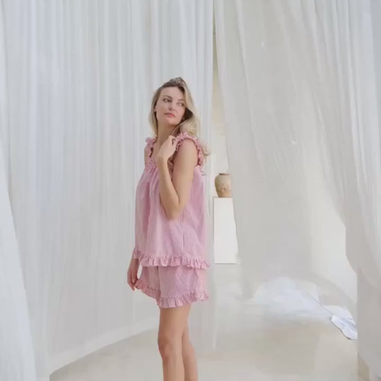 Video of Woman wearing a sleep set red and white pin stripe with ruffles on sleeves and hem of top and shorts. She is standing in front of a long white curtain.