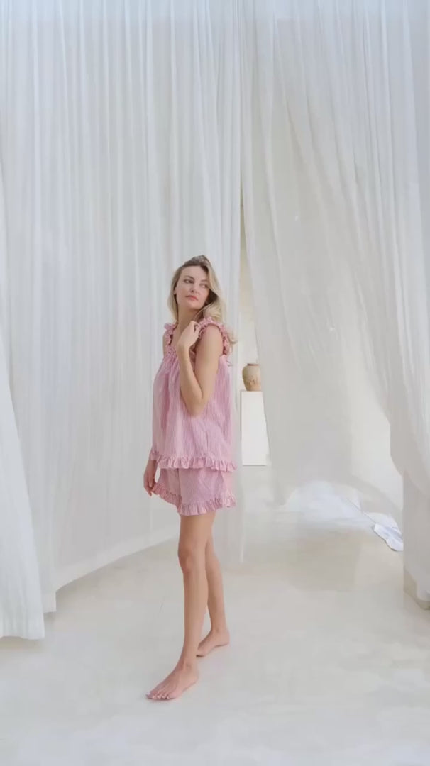 Video of Woman wearing a sleep set red and white pin stripe with ruffles on sleeves and hem of top and shorts. She is standing in front of a long white curtain.