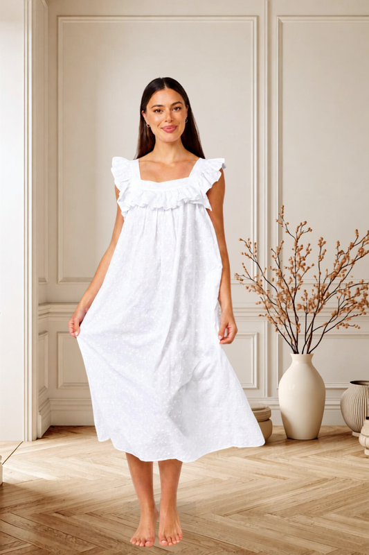 A woman wearing a white ruffled nightgown or midi dress stands barefoot in a bright, elegant room with paneled walls and a wood floor. She is holding out one side of the dress to show its drape.